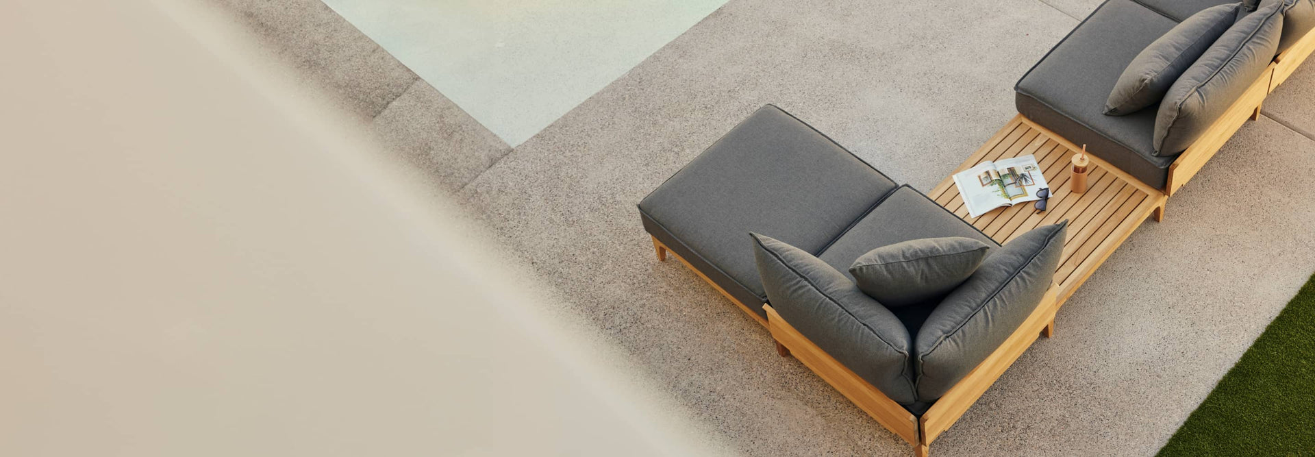 Teak outdoor chaise lounge chair with billowy gray cushions alongside a pool on a patio, photographed from above.