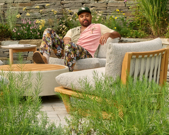 Hilton Carter lounging on a cushioned teak outdoor sectional in his Baltimore backyard, with a stone retaining wall and tall grasses behind him.