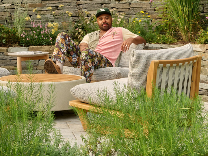 Hilton Carter lounging on a cushioned teak outdoor sectional in his Baltimore backyard, with a stone retaining wall and tall grasses behind him.