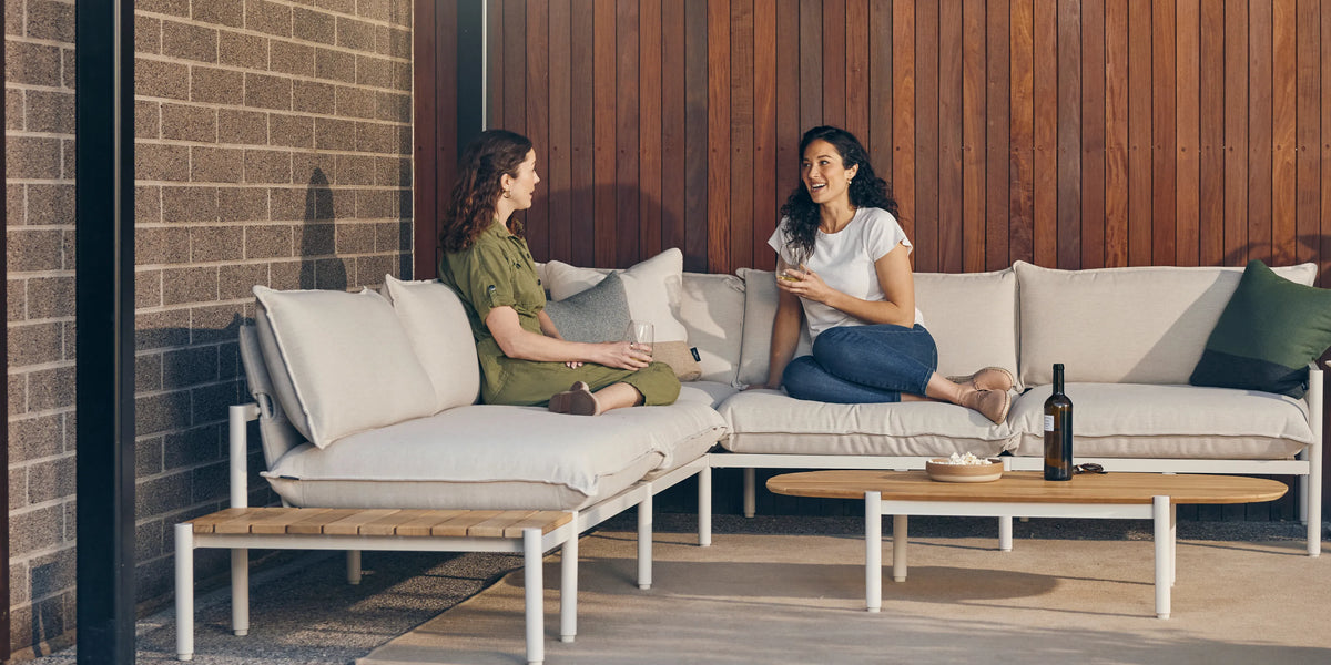 Two women talking while seated on an outdoor sectional with a teak end table