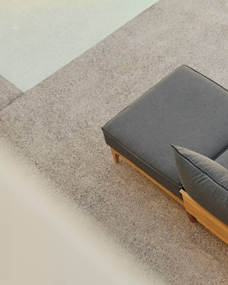 Teak outdoor chaise lounge chair with billowy gray cushions alongside a pool on a patio, photographed from above.