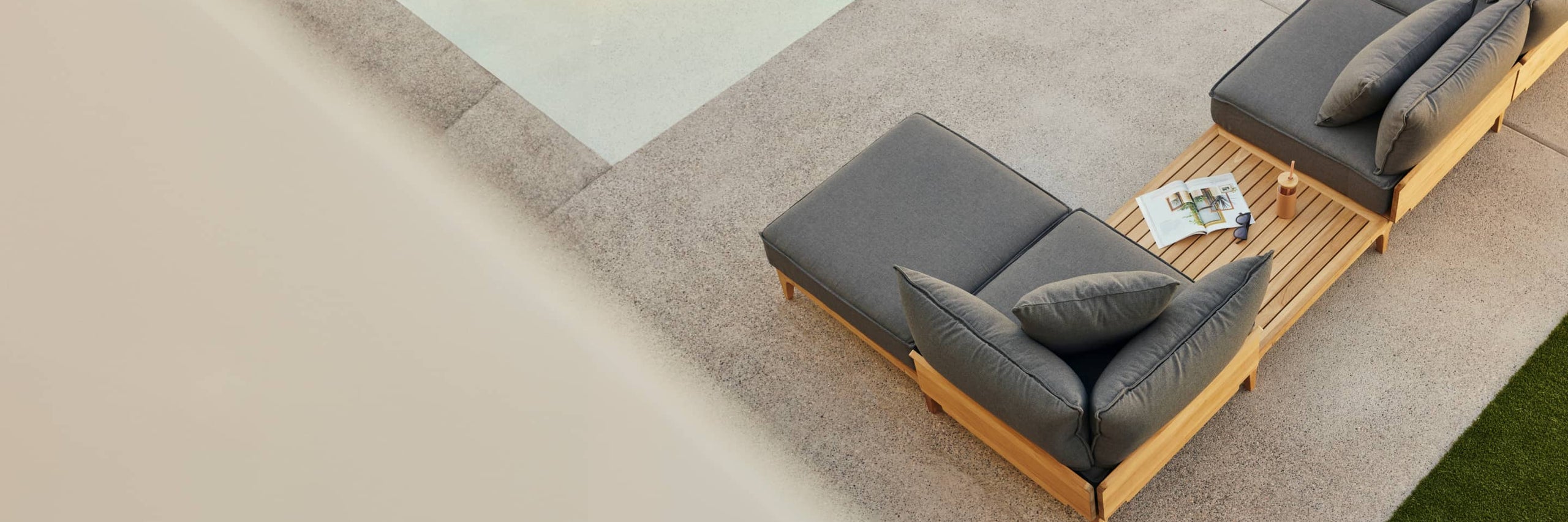 Teak outdoor chaise lounge chair with billowy gray cushions alongside a pool on a patio, photographed from above.