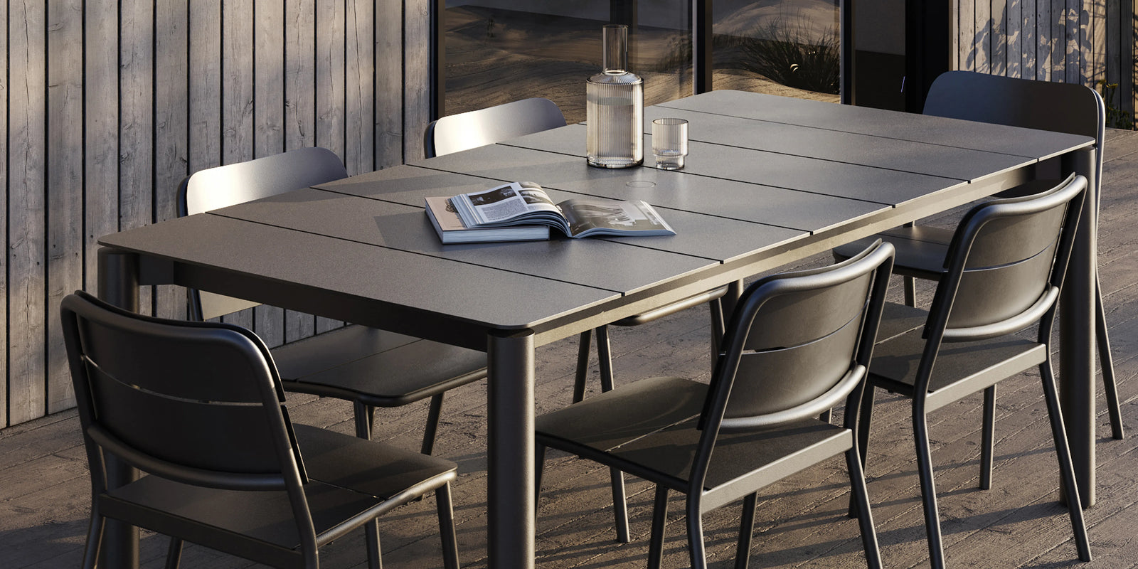 Neighbor Bandwidth Dining Table in Charcoal with chairs on a weathered wood deck, wood wall backdrop