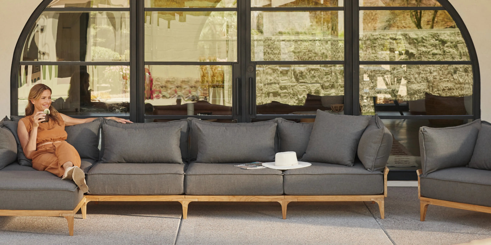 Person reclining on Neighbor Bluff Chaise Sectional in charcoal fabric with teak legs on a shaded patio