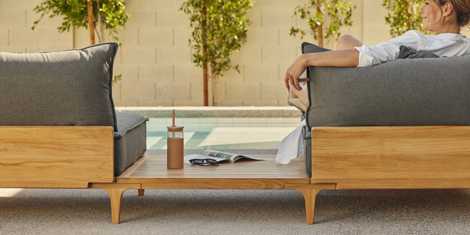 Person seated on Neighbor Bluff Sofa with End Table in charcoal and teak on patio, magazine and bottle on table