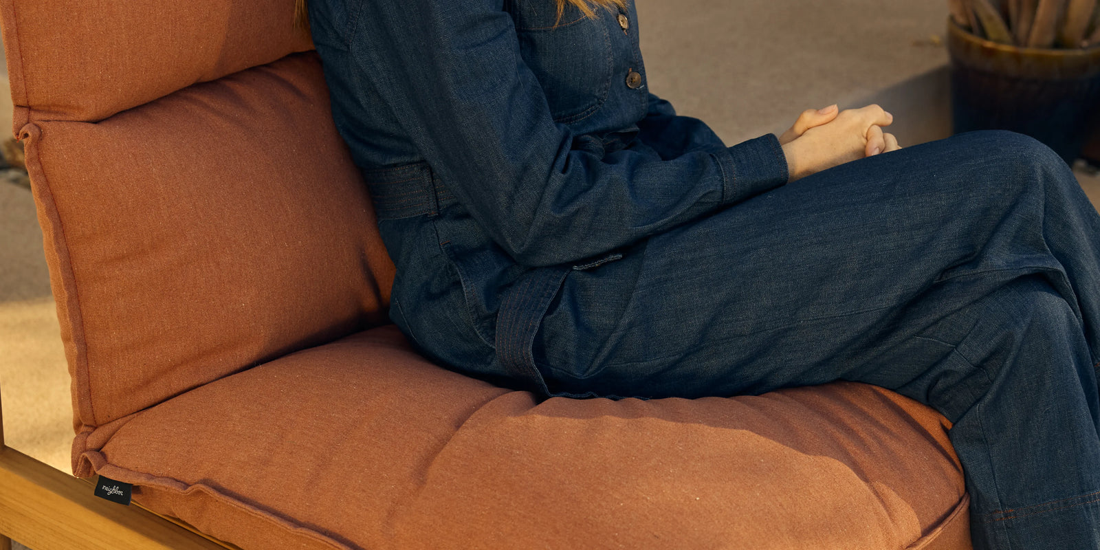 Woman seated in Neighbor Felloe Chair with rust cushion and teak frame on a sunlit patio, side table beside