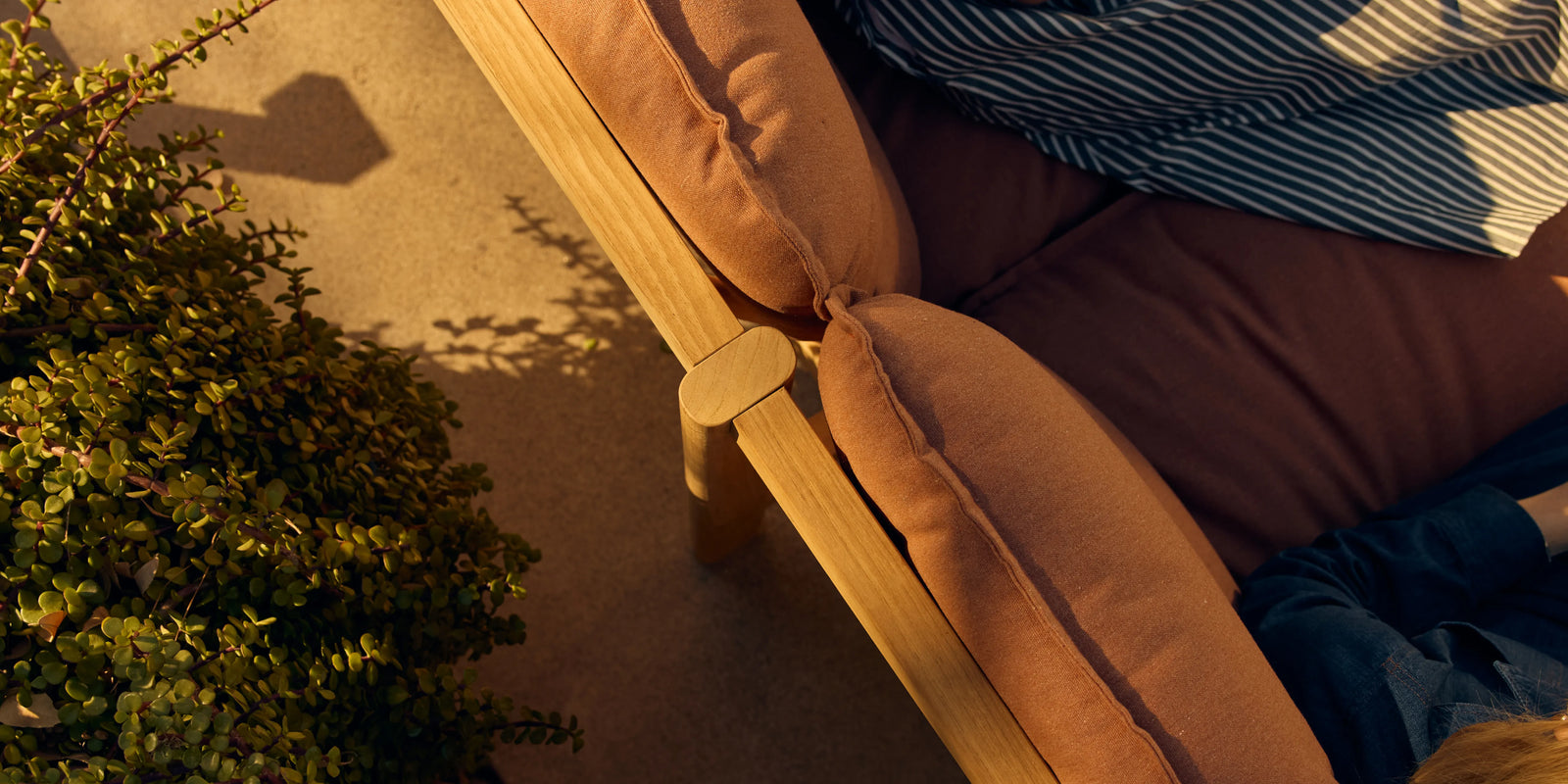 Overhead view of person relaxing on Neighbor Felloe Sofa with rust cushions and teak frame on sunny patio with greenery