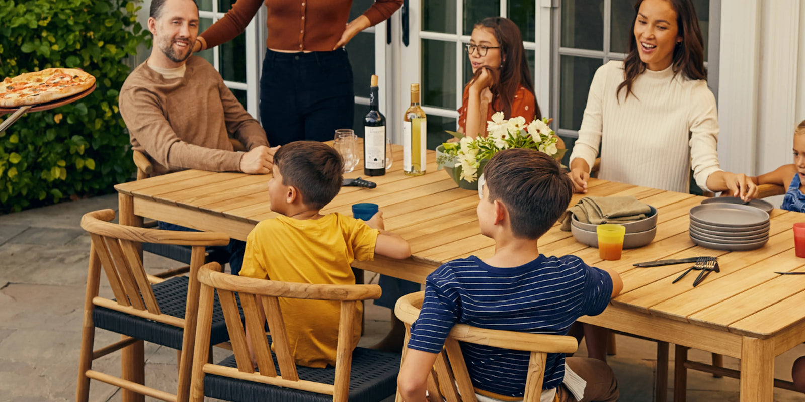 Neighbor Haven Dining Table in teak with woven-seat chairs on a covered porch, set with place settings