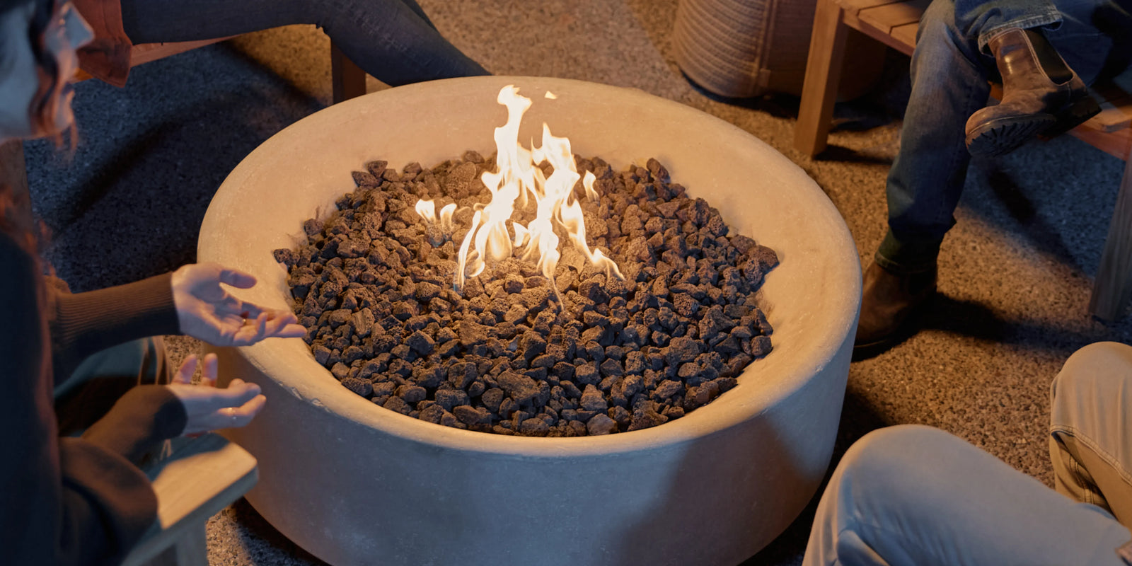 Neighbor Rook Round fire table with open flame on a gravel patio with Adirondack chairs at dusk