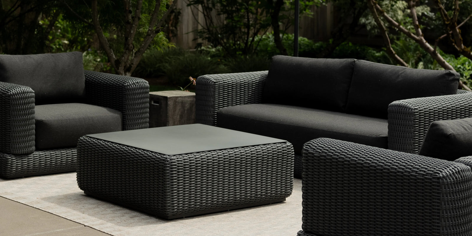 Neighbor Roundabout Sofa and chairs in dark wicker with charcoal cushions on a patio at dusk with garden backdrop