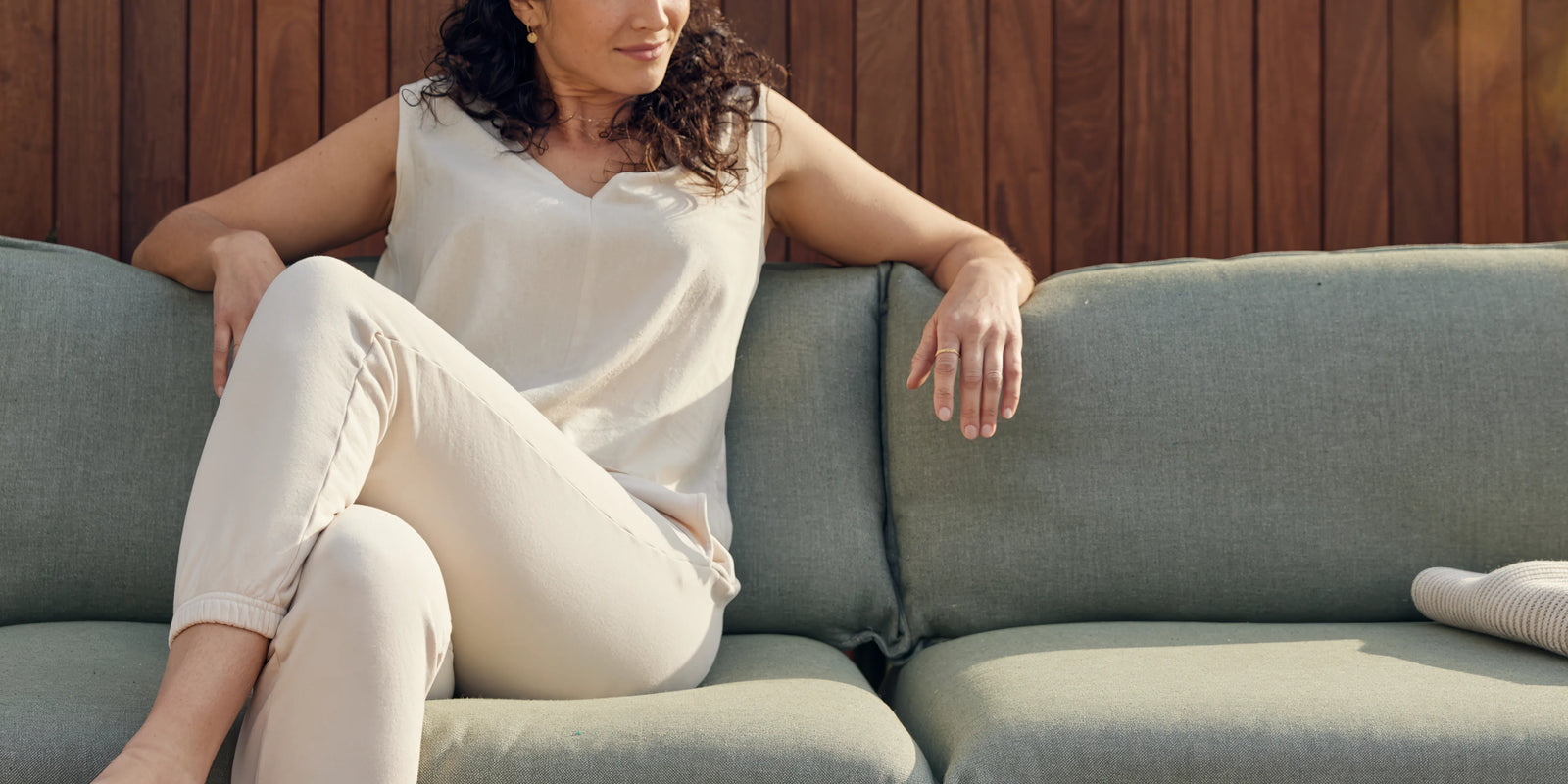 Woman relaxing on Neighbor Terra Sofa in sage green fabric with teak frame on a covered patio, wood-slat wall behind