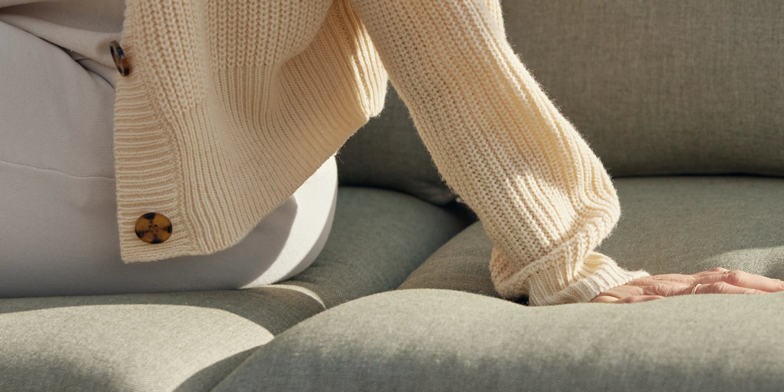 Closeup of person's legs resting on Neighbor Terra Sofa cushion in sage green fabric with knit throw in warm light