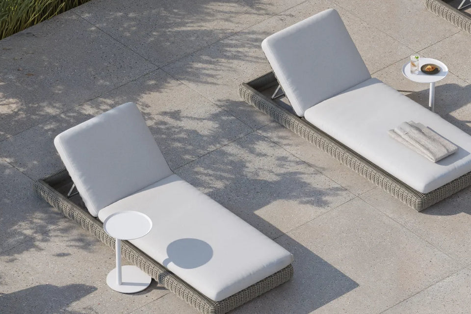 Top-down banner of four Neighbor Roundabout Sun Loungers in resin wicker with cream cushions arranged on a sunlit stone-tile patio, with a small round white side table holding a coffee and pastry between two of them; tall ornamental grasses cast soft shadows along the left edge.