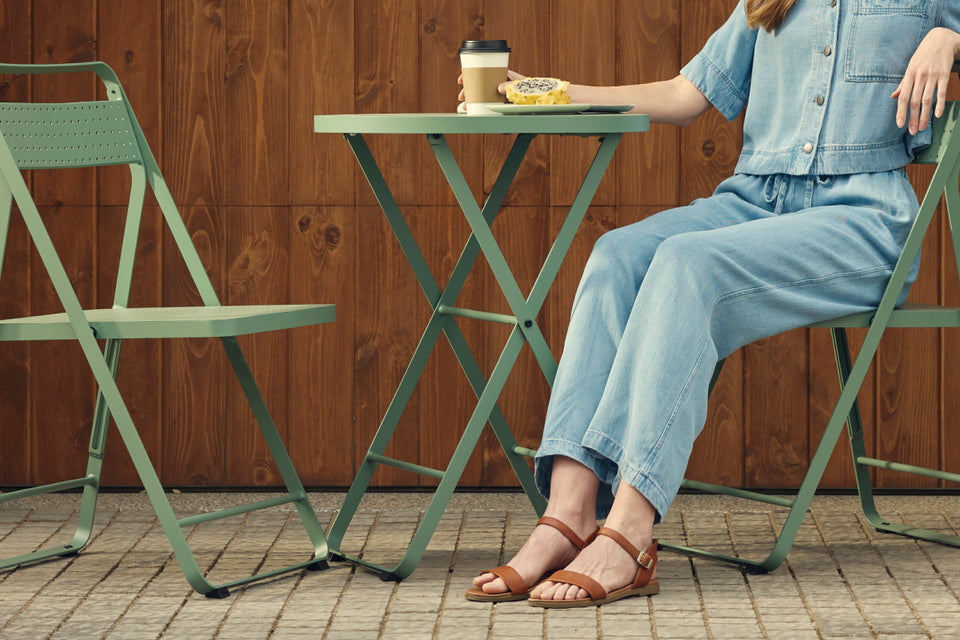 Wide banner of a woman in a denim jumpsuit and tan sandals seated on a sage-green Neighbor Take Two Bistro Chair on a brick-paver patio in front of a stained-wood wall, with a matching empty bistro chair beside the perforated steel bistro table that holds a coffee cup and pastry.