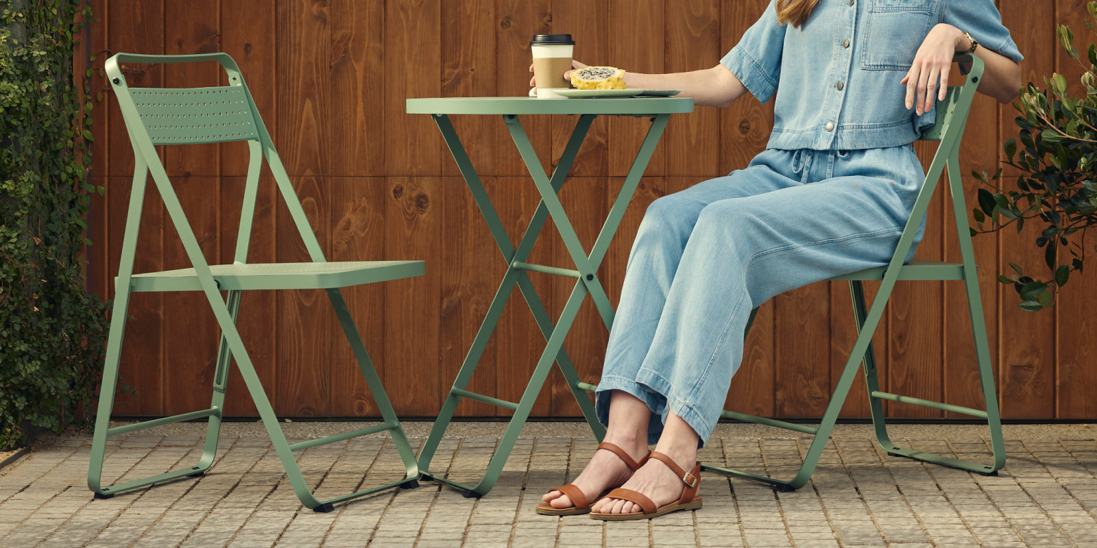 Wide banner of a woman in a denim jumpsuit and tan sandals seated on a sage-green Neighbor Take Two Bistro Chair on a brick-paver patio in front of a stained-wood wall, with a matching empty bistro chair beside the perforated steel bistro table that holds a coffee cup and pastry.