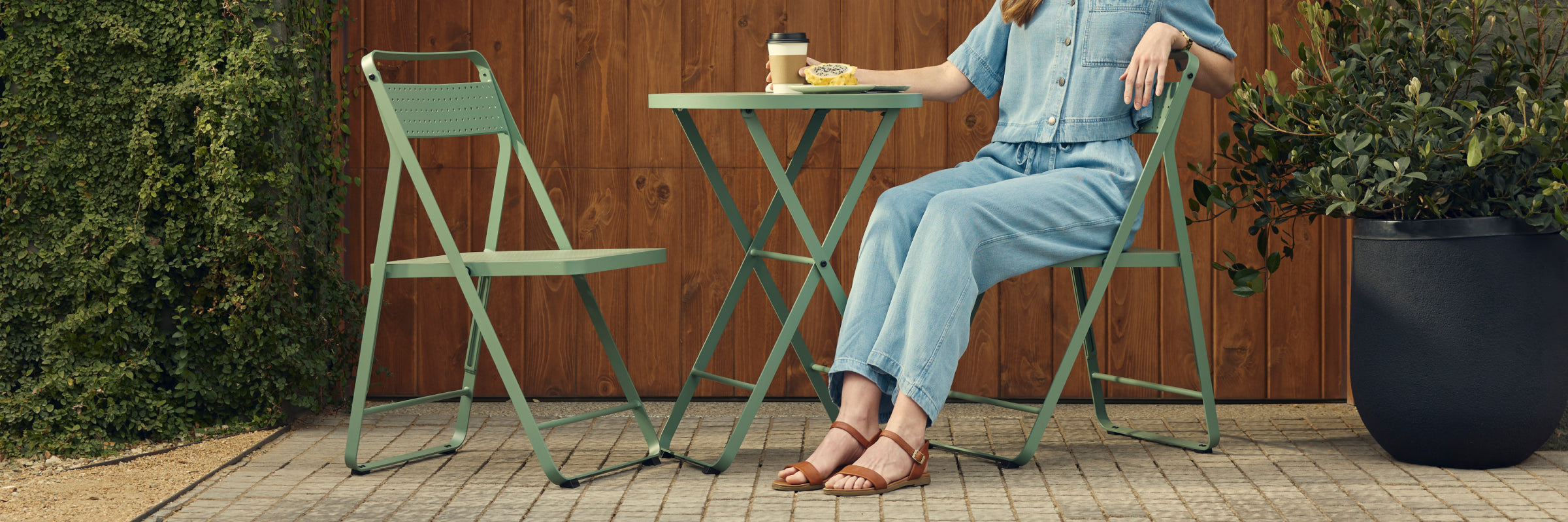 Wide banner of a woman in a denim jumpsuit and tan sandals seated on a sage-green Neighbor Take Two Bistro Chair on a brick-paver patio in front of a stained-wood wall, with a matching empty bistro chair beside the perforated steel bistro table that holds a coffee cup and pastry.