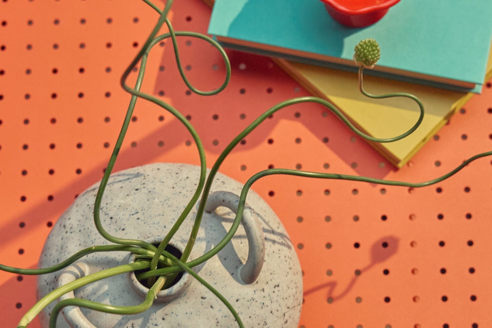 Tight overhead banner of the coral-red perforated steel top of a Neighbor Take Two Coffee Table styled with a stack of teal and yellow hardcover books, a small red candle, and a speckled stoneware vase holding a curling green vine.