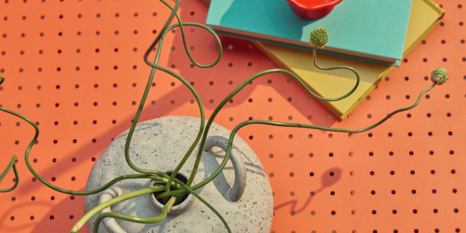 Tight overhead banner of the coral-red perforated steel top of a Neighbor Take Two Coffee Table styled with a stack of teal and yellow hardcover books, a small red candle, and a speckled stoneware vase holding a curling green vine.