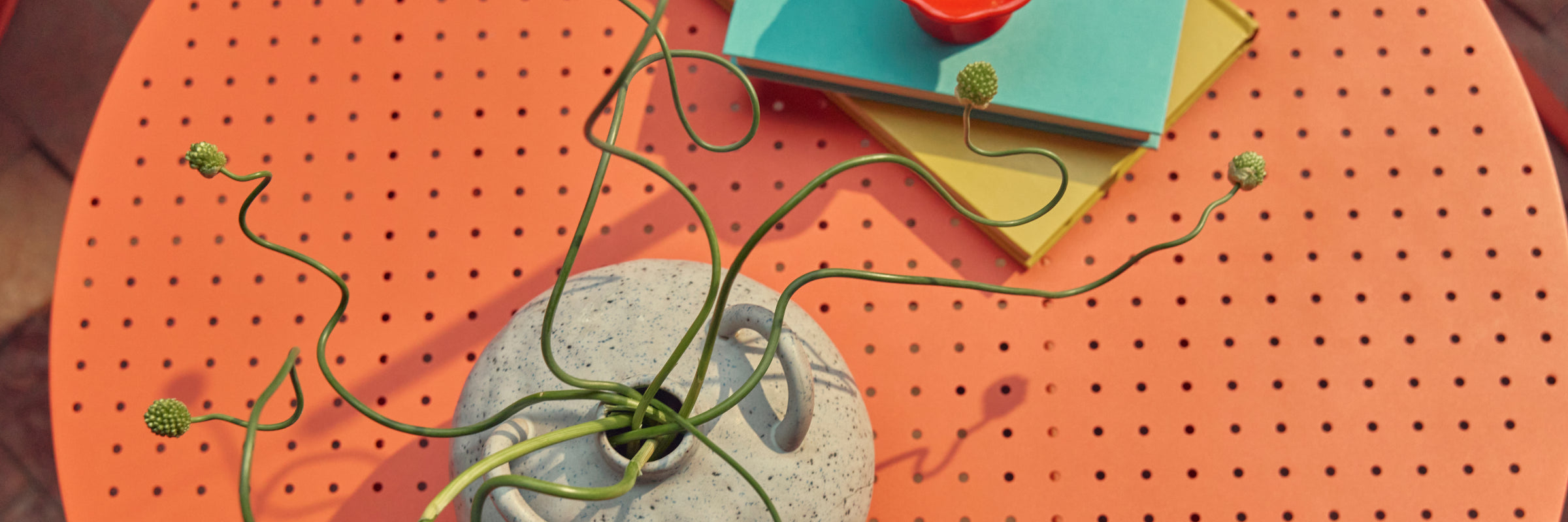 Tight overhead banner of the coral-red perforated steel top of a Neighbor Take Two Coffee Table styled with a stack of teal and yellow hardcover books, a small red candle, and a speckled stoneware vase holding a curling green vine.