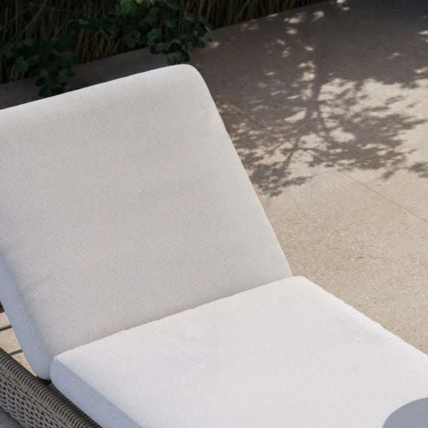 Elevated three-quarter view of a Neighbor Roundabout Sun Lounger in taupe-gray resin wicker with cream all-weather cushion on a sunlit stone-tile patio, with dappled tree shadows and tropical foliage behind it and a small white side table holding lemon water and a croissant alongside.
