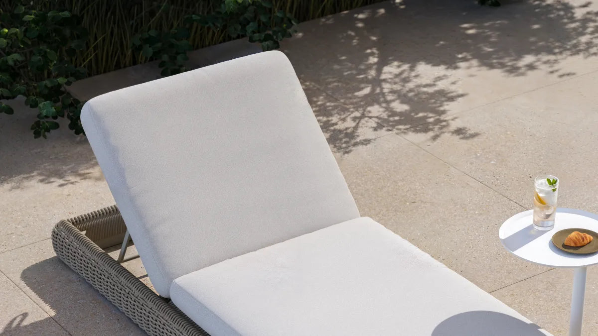 Elevated three-quarter view of a Neighbor Roundabout Sun Lounger in taupe-gray resin wicker with cream all-weather cushion on a sunlit stone-tile patio, with dappled tree shadows and tropical foliage behind it and a small white side table holding lemon water and a croissant alongside.