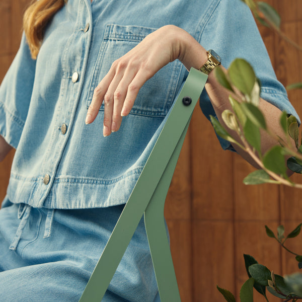 Cropped lifestyle shot of a woman in a denim shirt and pants seated on a sage-green Neighbor Take Two Bistro Chair, hand resting on the chair's slim folding-steel backrest, in front of a stained-wood fence with glossy laurel foliage to the side.