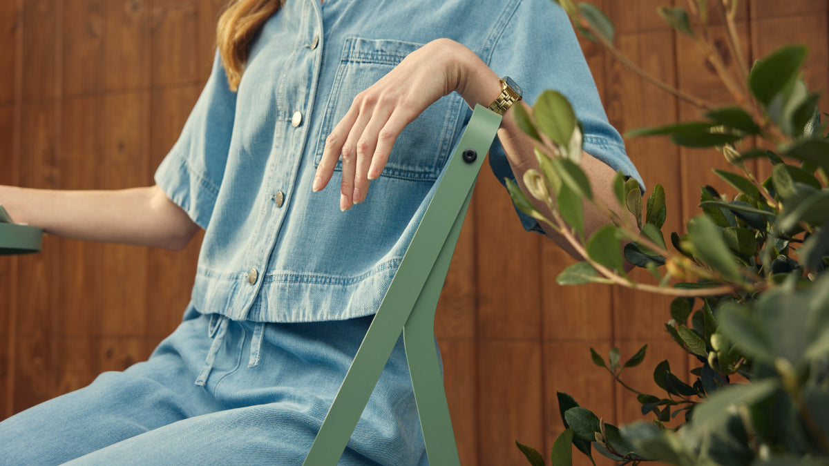 Cropped lifestyle shot of a woman in a denim shirt and pants seated on a sage-green Neighbor Take Two Bistro Chair, hand resting on the chair's slim folding-steel backrest, in front of a stained-wood fence with glossy laurel foliage to the side.