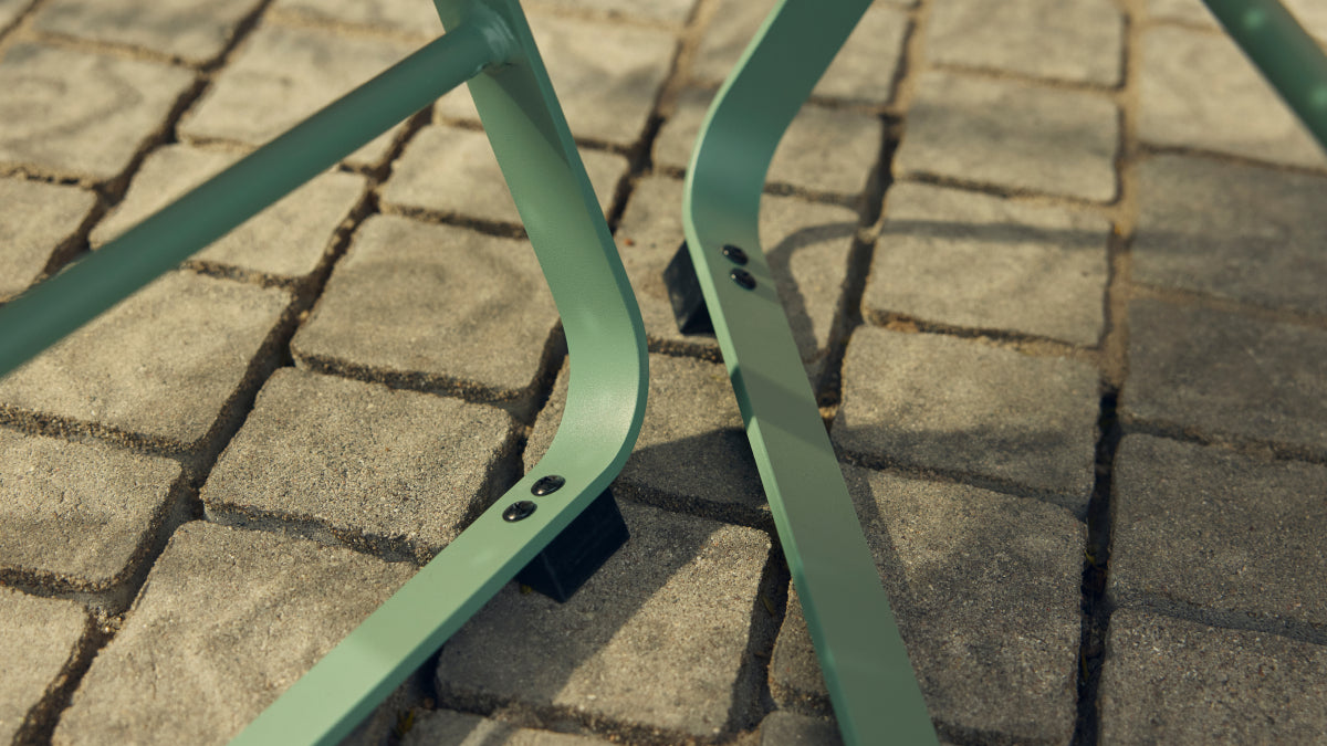Tight ground-level detail of two sage-green powder-coated steel chair legs from a Neighbor Take Two Bistro Chair planted on weathered cobblestone pavers, showing the protective black foot caps where the legs meet the stone.
