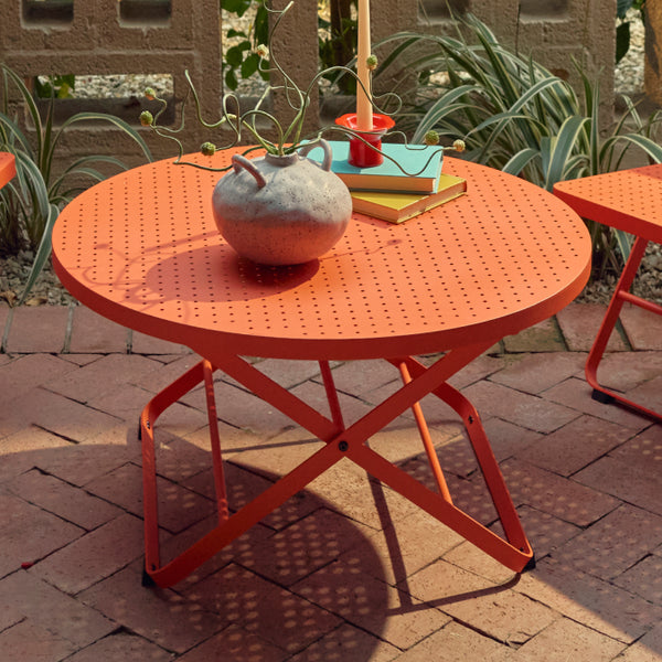 Three-quarter view of a coral-red Neighbor Take Two Coffee Table with its round perforated steel top centered between two matching coral-red Lounge Chairs on a red-brick paver patio, with a textured stoneware vase, stacked books and a candle styled on top.