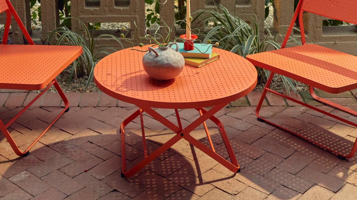 Three-quarter view of a coral-red Neighbor Take Two Coffee Table with its round perforated steel top centered between two matching coral-red Lounge Chairs on a red-brick paver patio, with a textured stoneware vase, stacked books and a candle styled on top.