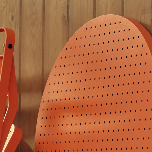 Tight detail of a coral-red Neighbor Take Two Coffee Table with its round perforated steel top folded down and leaned against a stained-wood plank wall, with a coral-red folding chair partially in frame to the left.