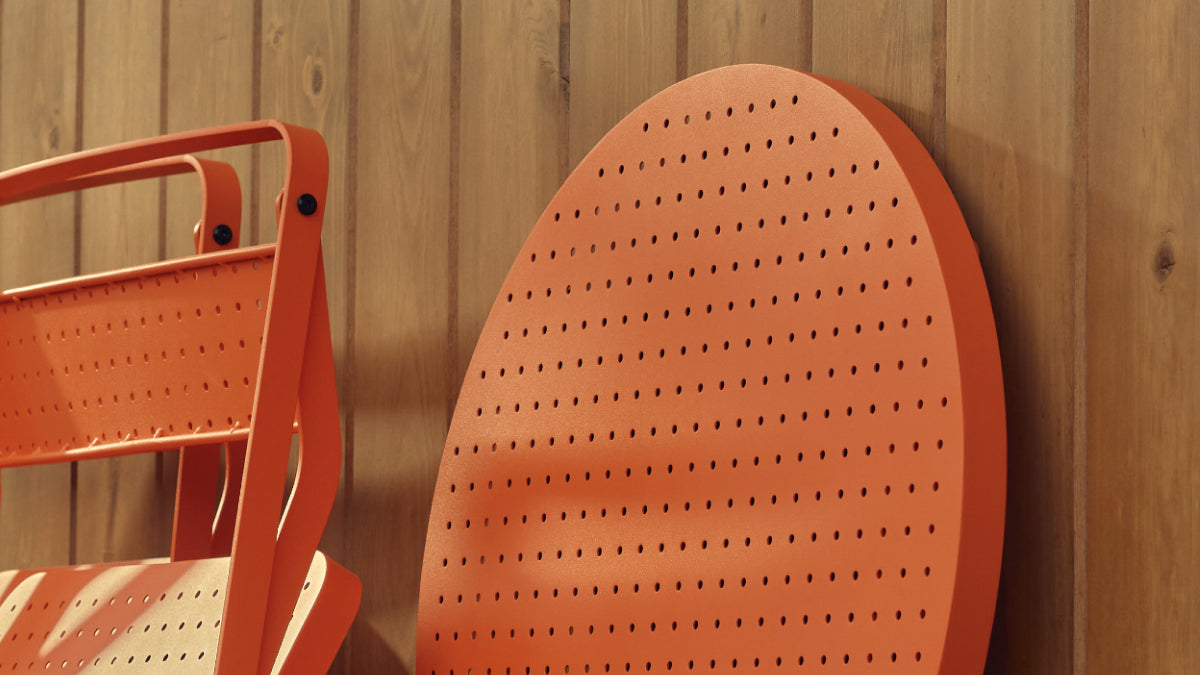 Tight detail of a coral-red Neighbor Take Two Coffee Table with its round perforated steel top folded down and leaned against a stained-wood plank wall, with a coral-red folding chair partially in frame to the left.