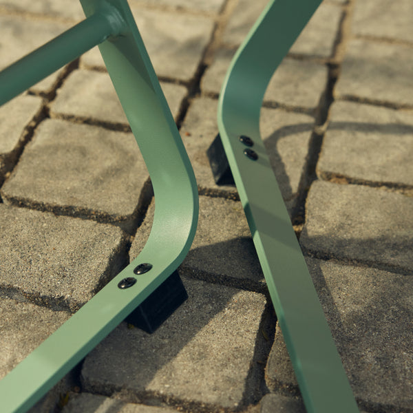 Tight ground-level detail of two sage-green powder-coated steel legs of a Neighbor Take Two Coffee Table with their black rubber foot caps planted firmly on weathered cobblestone pavers.