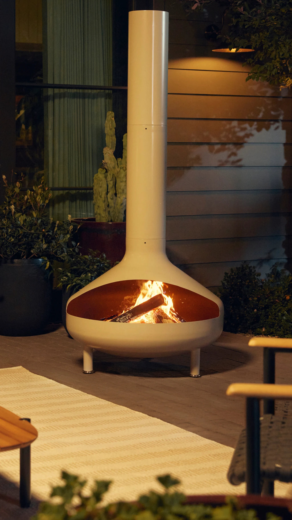 Modern chiminea made of metal with a tall flue with a fire burning inside set in outdoor patio area.
