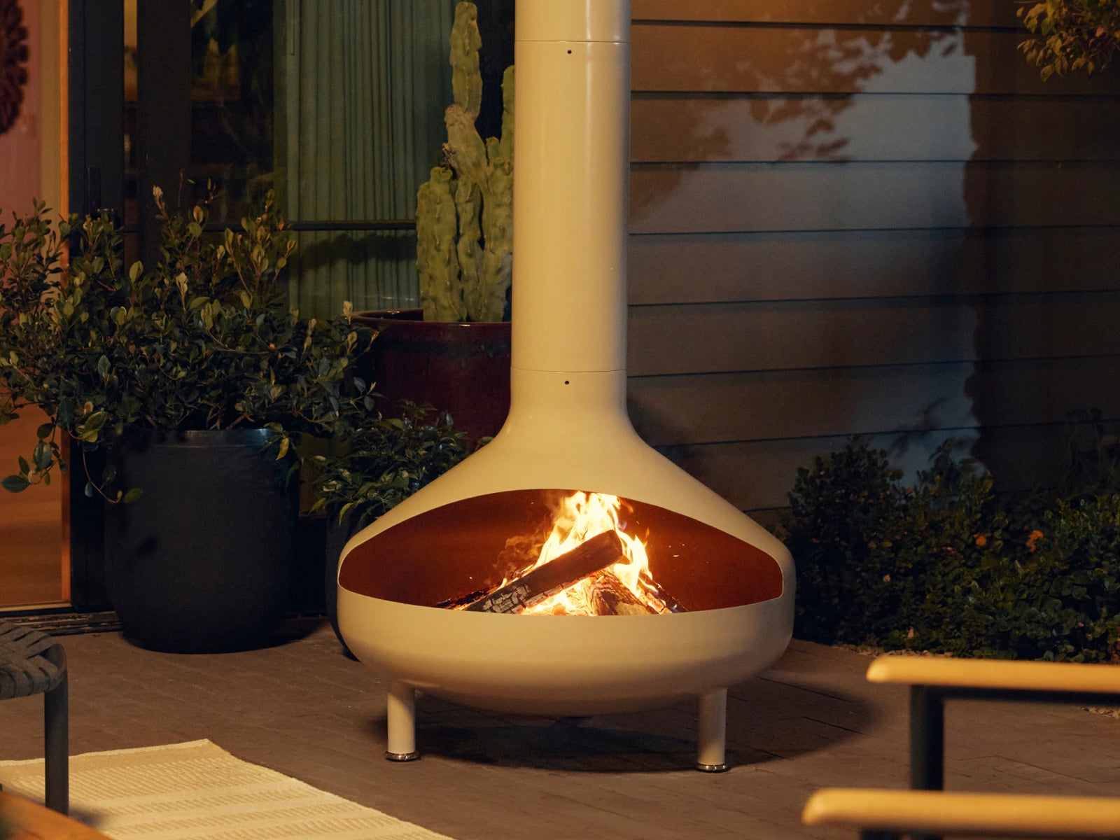 Modern chiminea made of metal with a tall flue with a fire burning inside set in outdoor patio area.