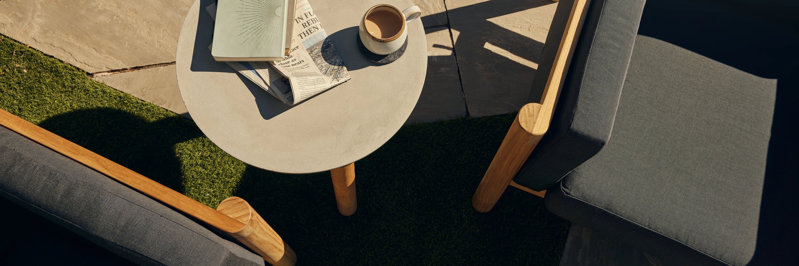Overhead shot of a round concrete side table with a book and coffee cup, flanked by teak lounge chairs on a lawn