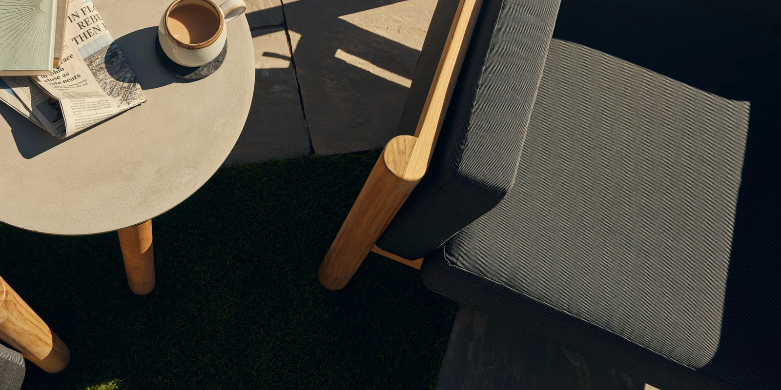 Overhead shot of a round concrete side table with a book and coffee cup, flanked by teak lounge chairs on a lawn
