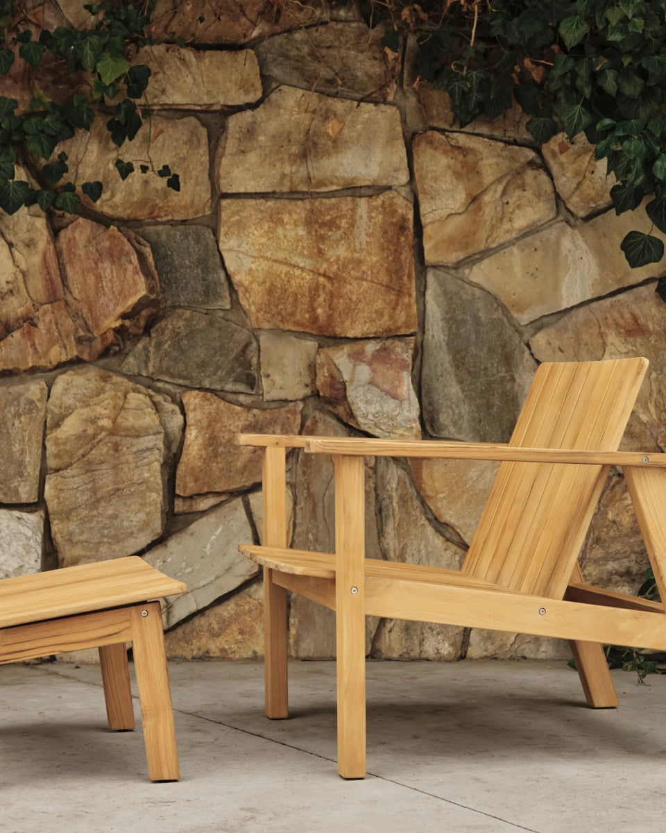 Teak adirondack-style lounge chair and matching footstool against a rustic stone wall