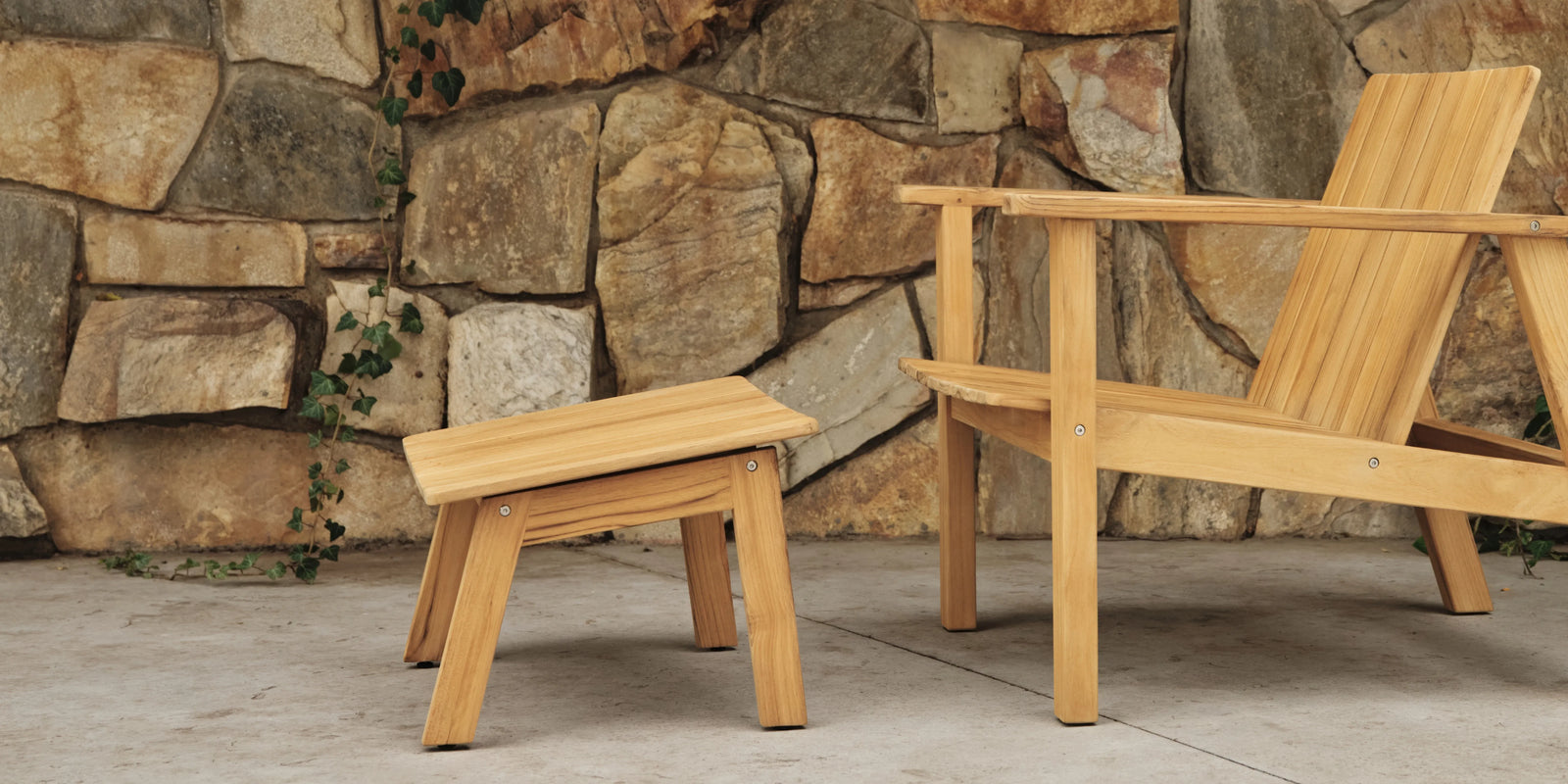 Teak adirondack-style lounge chair and matching footstool against a rustic stone wall