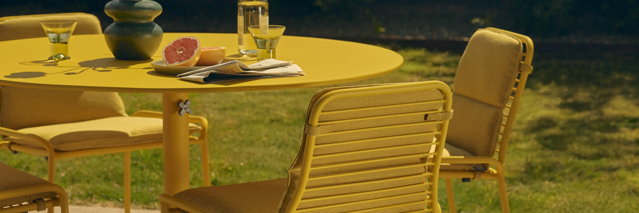 Yellow round aluminum dining table and matching slatted chairs on a lawn, styled with grapefruit and drinks