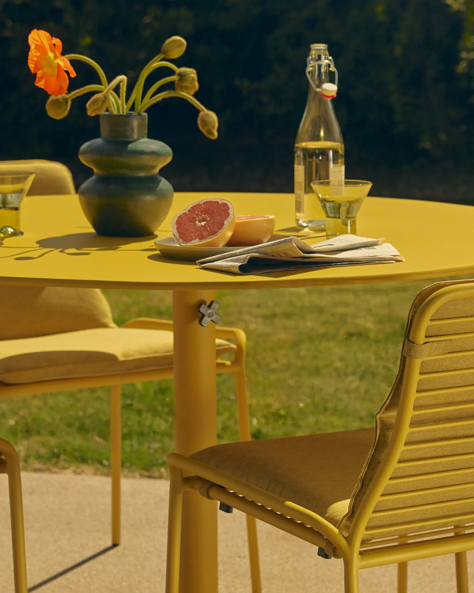 Yellow round aluminum dining table and matching slatted chairs on a lawn, styled with grapefruit and drinks