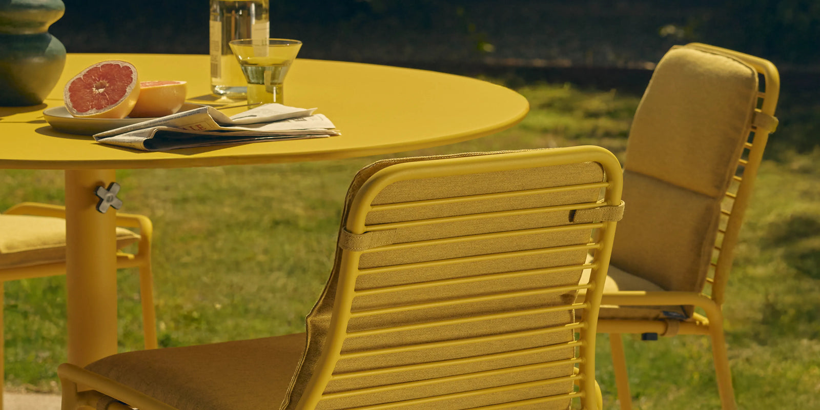 Yellow round aluminum dining table and matching slatted chairs on a lawn, styled with grapefruit and drinks