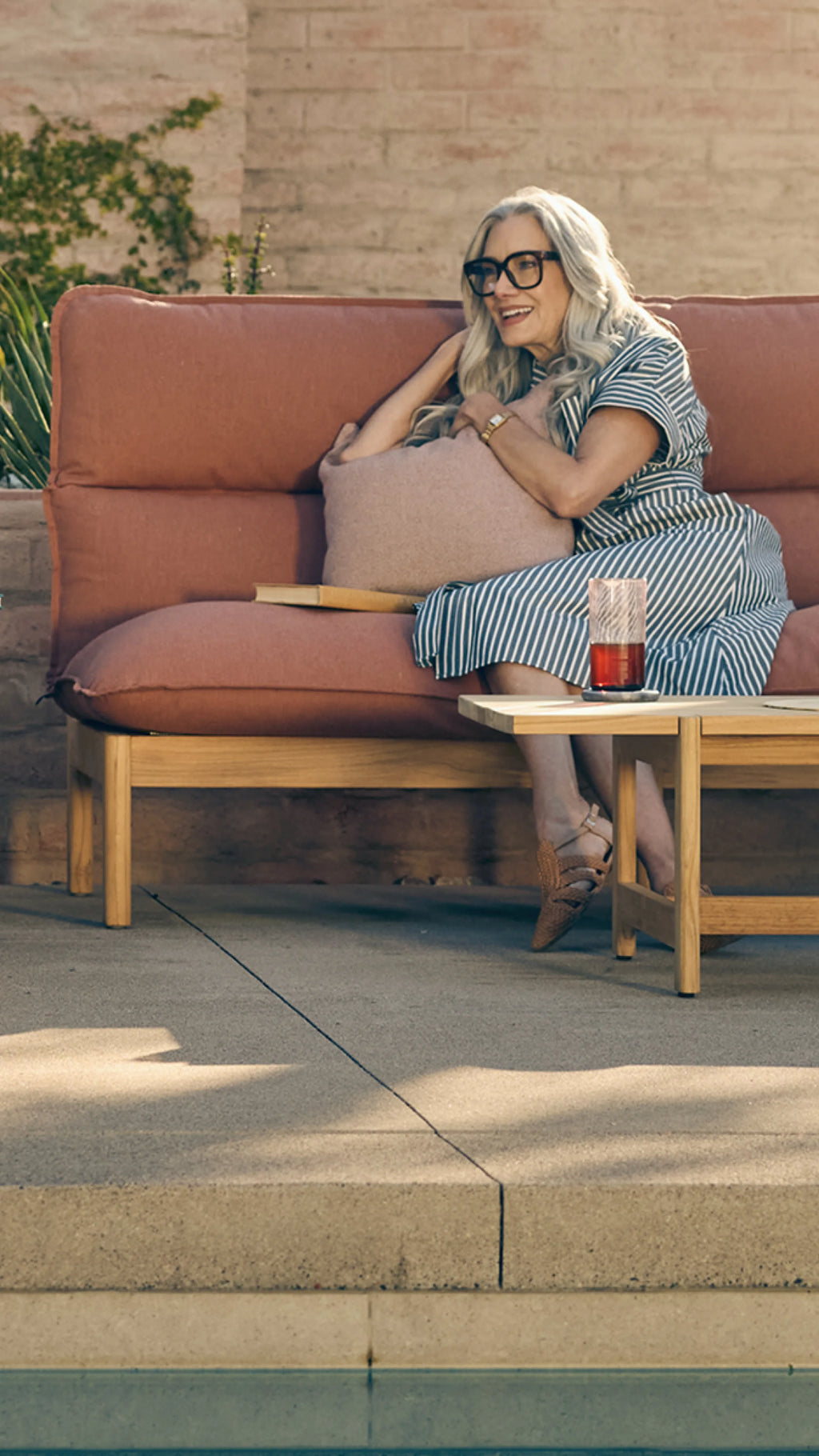 Woman relaxing on a teak outdoor sofa with terracotta cushions beside a pool