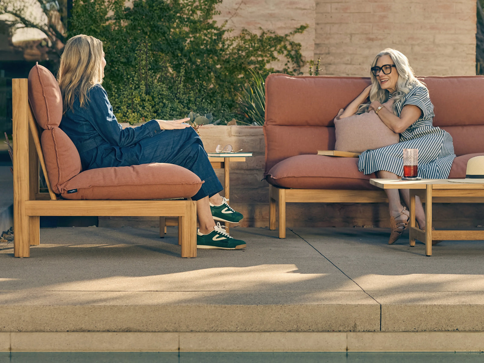 Two women relaxing poolside on teak outdoor sofa and lounge chair with terracotta cushions