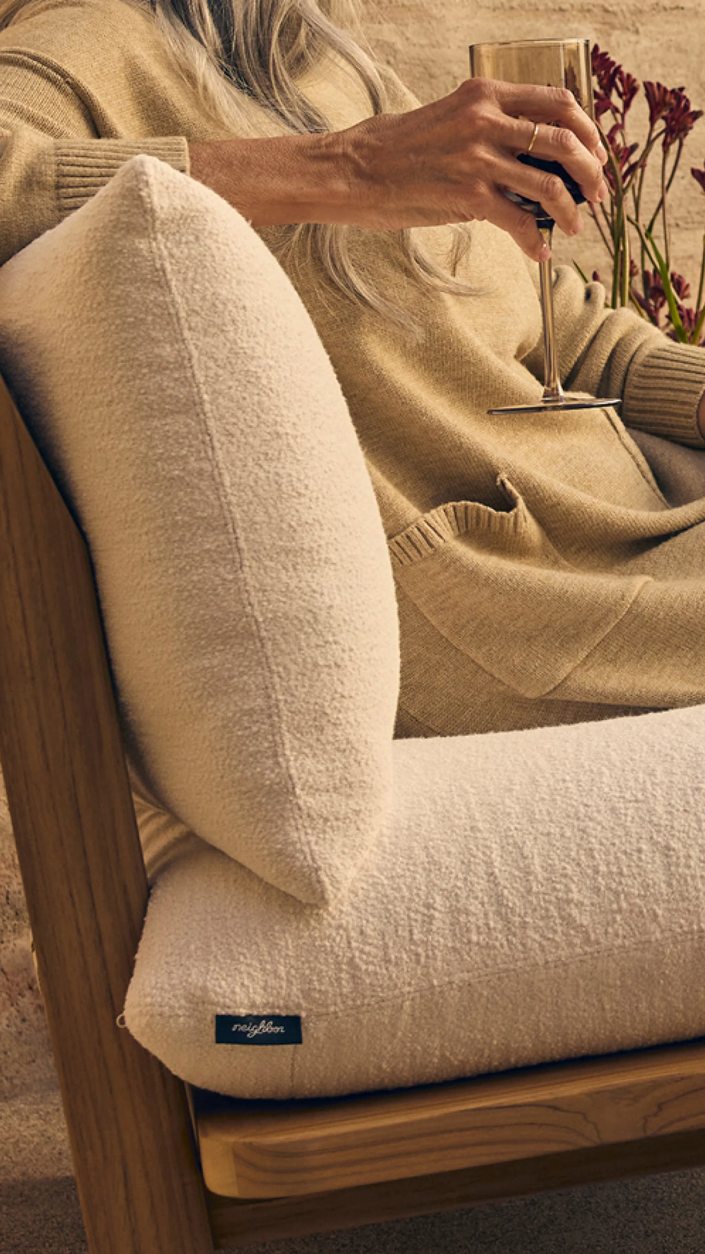 Close-up of a woman in a teak lounge chair with cream boucle cushions and Neighbor label, holding a glass