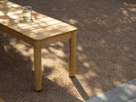 Neighbor Haven 72-inch Bench with natural finish wood and glass of water on surface, on a gravel patio with tree shadows