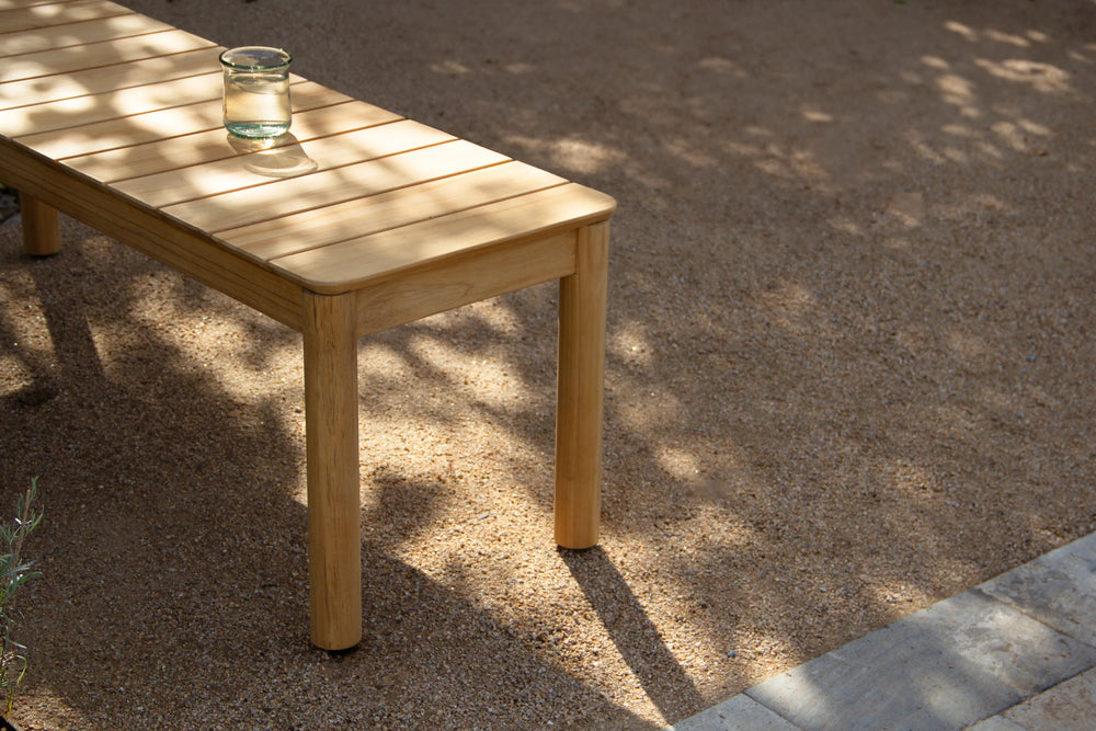 Neighbor Haven 72-inch Bench with natural finish wood and glass of water on surface, on a gravel patio with tree shadows