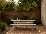 A light-colored outdoor picnic table with two matching benches on a paved patio, set against a wooden fence and greenery in a backyard setting.