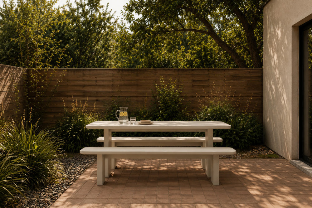 A light-colored outdoor picnic table with two matching benches on a paved patio, set against a wooden fence and greenery in a backyard setting.