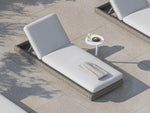 Top-down lifestyle shot of a Neighbor Roundabout Sun Lounger in taupe-gray resin wicker with thick cream cushion on a sunlit stone-tile poolside patio, with a folded towel set on the seat and a small white side table holding lemon water and a croissant beside it.